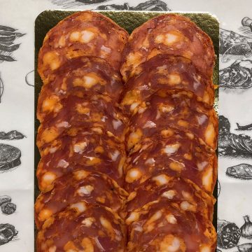 A tray of thinly sliced cured meat, likely chorizo, arranged neatly in overlapping rows on a gold-colored board with illustrated meat drawings and a "Cobble Lane Cured" label in the background.