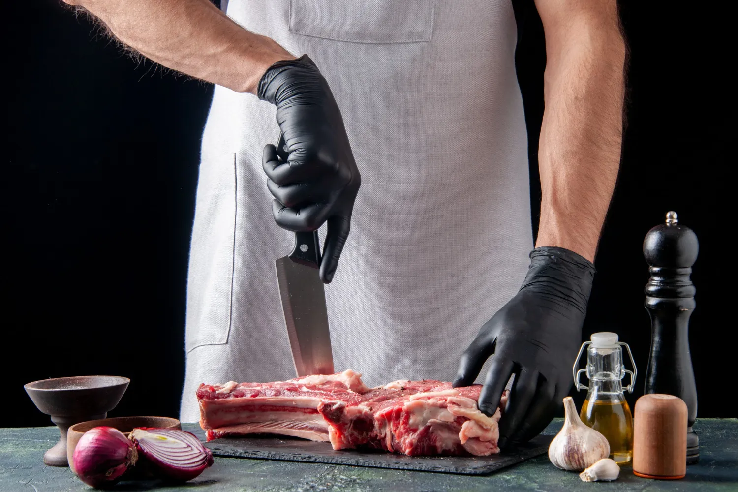 A person in a white apron and black gloves cuts raw meat on a black cutting board, surrounded by onion, garlic, oil, a pepper grinder, and salt—showcasing ingredients sourced through Regenerative Farming for Foodservice.