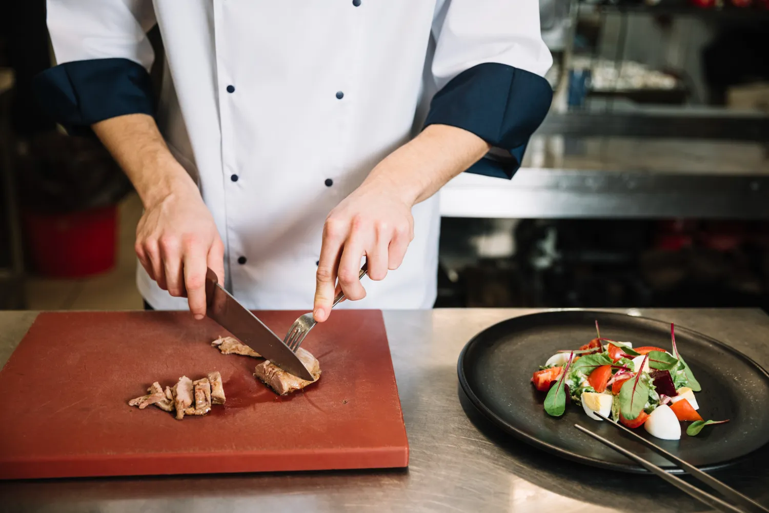 A chef in a white coat slices cooked meat on a red cutting board in a kitchen focused on Regenerative Farming for Foodservice, with a plate of colorful salad on the counter nearby.